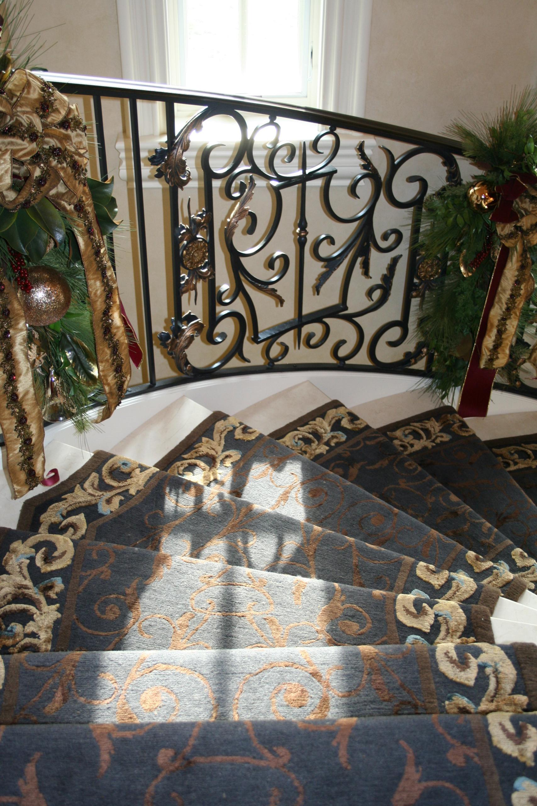 A curving staircase featuring patterned blue and gold runner rugs and an ornate wrought-iron railing decorated with holiday greenery and ribbons.
